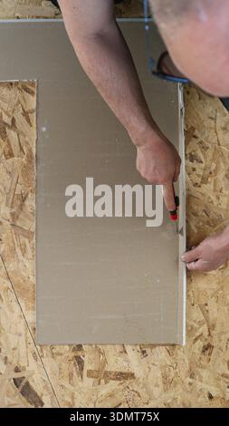 A utility knife in the hands of a repairman sitting on the floor and cutting a sheet of cardboard or plywood in a straight line, on the floor Stock Photo