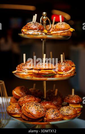 Small sesame seed burgers are arranged on a two-tier metal stand. The warm light highlights the buns, cheese, and fillings. Stock Photo