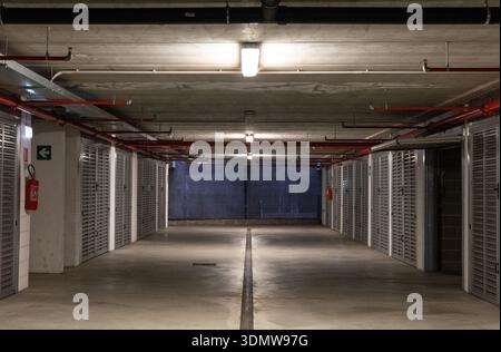 Empty underground parking garage corridor with concrete ceiling, industrial lighting and metal storage doors, minimalist urban interior. Stock Photo