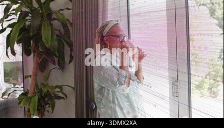 Sipping clear glass, senior woman wearing light robe and glasses, standing at sliding door at home Stock Photo