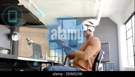 Interacting man wearing tan sweater white VR headset and headphones using tablet hologram at office Stock Photo