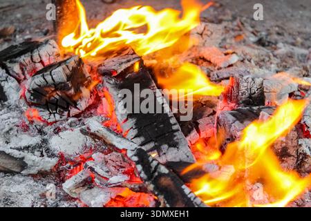 Glowing red-hot embers and burning wood logs in an active fire. Flames swirl around the chunks of firewood, creating a high-contrast scene. Stock Photo
