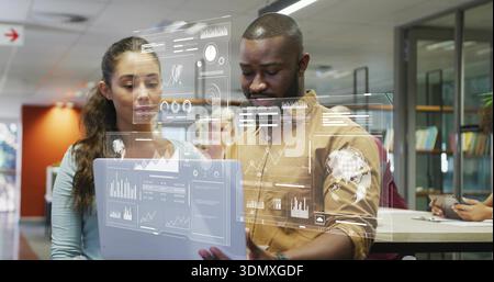 Reviewing two coworkers in tan shirt and blue top studying laptop at office with holographic charts Stock Photo