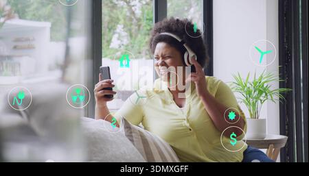 Using phone, woman in yellow shirt and white headset touching earcup on couch with AR icons Stock Photo