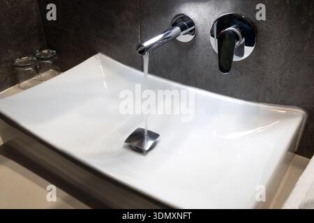 Closeup of running water from a wall mounted chrome faucet into a white rectangular ceramic basin in a contemporary bathroom with dark tiles and soft lighting Stock Photo