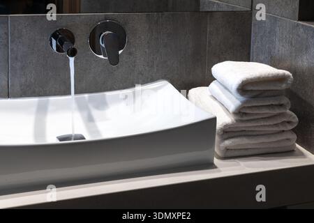 White rectangular basin with wall mounted chrome mixer running water beside stacked soft towels in a contemporary hotel style bathroom with dark stone tiles and clean lighting Stock Photo