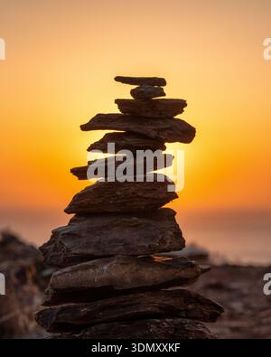 Balanced stack of smooth stones forming a zen cairn on a rocky coastline during golden sunset with soft pastel sky and calm sea, representing stabilit Stock Photo