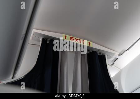 Closeup of an airplane cabin with curtains dividing classes and a red EXIT sign above the aisle, showing passenger safety signage inside a modern commercial aircraft interior Stock Photo