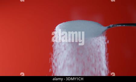 Sugar is poured from a spoon with a red background showing falling granules and a focused view of the sugar Stock Photo