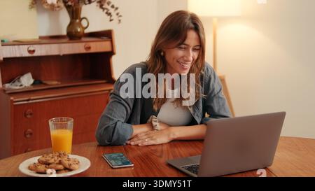 A cheerful woman sits at a wooden table with a laptop, a glass of orange juice, and cookies, enjoying a relaxed moment while working from home. Stock Photo