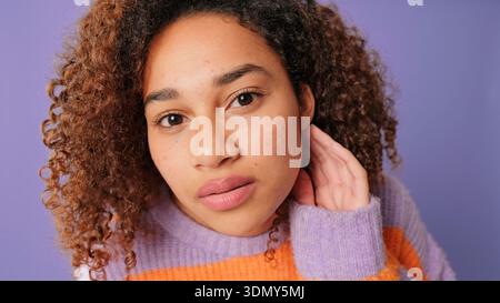 A close-up portrait of a young woman with curly hair, wearing a lavender sweater with orange stripes, against a purple background. She gazes softly at Stock Photo