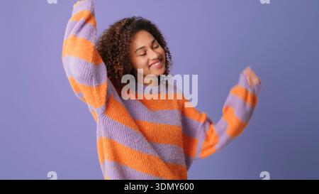 A smiling woman wearing a bold orange and lavender striped knit sweater dances with arms raised in a bright studio setting. Energetic, cheerful lifest Stock Photo
