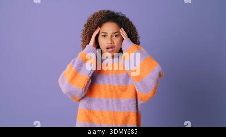 A young woman with curly hair wears a purple and orange striped sweater, posing against a purple backdrop. She holds her head with a surprised express Stock Photo