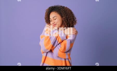 A warm, content woman wraps her arms around herself in a lavender and orange striped sweater, eyes closed with a gentle smile, against a soft purple b Stock Photo