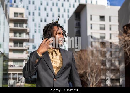 African American businessman with dreadlocks holds a phone while standing in a modern urban setting. He is dressed in a smart suit, looking confidentl Stock Photo