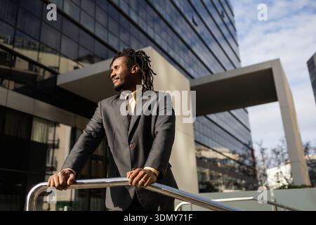 African American businessman with dreadlocks stands confidently outdoors, leaning on a railing, with a modern glass building backdrop, embodying profe Stock Photo