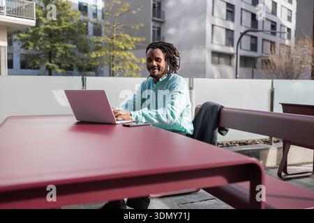 African American businessman with dreadlocks working on a laptop at an outdoor table. He is dressed in a casual green shirt and appears focused and en Stock Photo