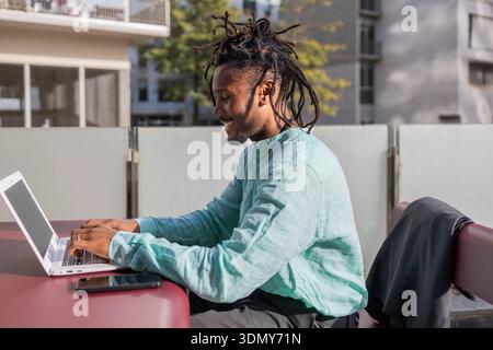 African American businessman with dreadlocks works on a laptop outdoors, showcasing productivity and modern work style. He's wearing a light green shi Stock Photo