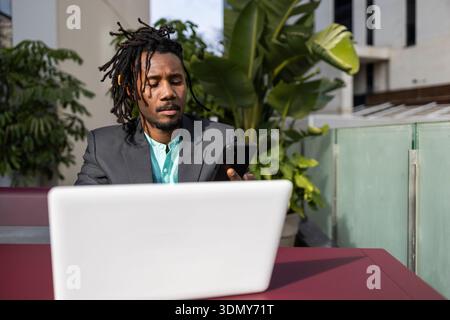 African American businessman with dreadlocks sits outdoors at a modern workspace, focused on his smartphone and laptop. Urban setting with greenery in Stock Photo