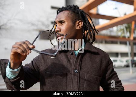 A confident African American businessman with dreadlocks, dressed in a brown coat, holds sunglasses. He stands outdoors against a blurry urban backdro Stock Photo