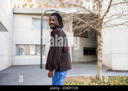 African American businessman with dreadlocks stands confidently in an urban setting, wearing a dark jacket and blue jeans. The scene exudes profession Stock Photo