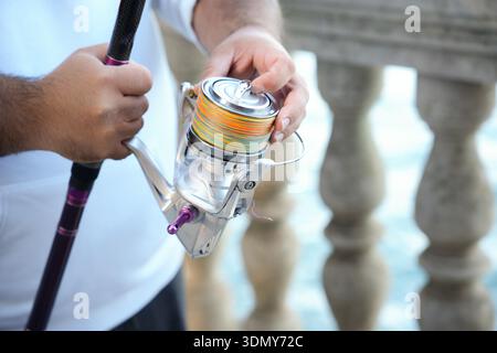 A close-up of a person's hand holding a fishing rod with a shiny, colorful reel. The background showcases a blurred stone railing and serene water, cr Stock Photo