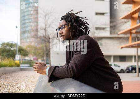 An African American businessman with dreadlocks and glasses, wearing a dark coat, leans on a railing outdoors, contemplating and observing the urban s Stock Photo