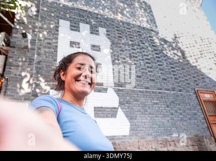 A woman smiles warmly while standing in front of a brick wall featuring artistic Chinese letters. The setting is Chongqing, creating a vibrant and cul Stock Photo