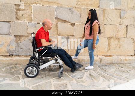 A joyful outdoor scene showing a man in a wheelchair interacting with a smiling young woman. The setting emphasizes inclusivity and barrier-free envir Stock Photo