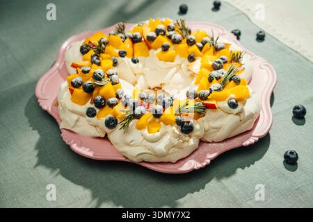 Delicious pavlova cake topped with fresh blueberries and peaches, garnished with sprigs of rosemary on a pink plate, creating a festive and elegant de Stock Photo