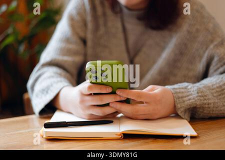 A teenager intently looks at a smartphone while sitting at a wooden table. An open notebook and pen are on the table, indicating a blend of technology Stock Photo