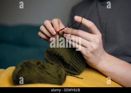 A close-up of a man knitting with olive green yarn, focusing on his hands and the knitting needles. The yarn ball and knitted fabric are also visible, Stock Photo