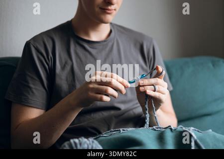 A young man in casual attire sits comfortably on a sofa, crocheting with blue yarn and a hook. His focus is on his craft, creating a serene and cozy a Stock Photo