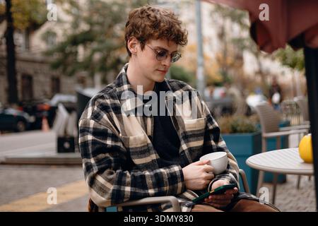 A young man sits comfortably at an outdoor cafe on a pleasant day, sipping coffee while using his smartphone. The serene atmosphere highlights his rel Stock Photo