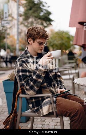 A young man sits outdoors at a cafe, enjoying a hot drink while browsing his smartphone. He's dressed in casual attire, with trees and street views in Stock Photo