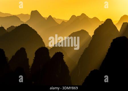 Golden light washes over layered karst limestone mountains in Yangshuo, Guangxi. Soft haze, deep silhouettes, and sun rays create a dramatic and atmos Stock Photo