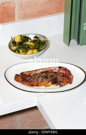 A juicy rib-eye steak is artfully presented on a white plate, accompanied by a bowl of roasted potatoes and peppers. Set in a cozy restaurant kitchen Stock Photo