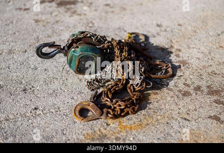 A coiled, rusty metal chain and hook lie on a rough concrete surface, bathed in sunlight. The image highlights the texture and detail of the aged mate Stock Photo