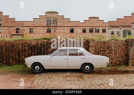 A vintage white sedan is parked parallel to a weathered red brick wall in front of an old industrial building. The setting evokes a nostalgic and hist Stock Photo