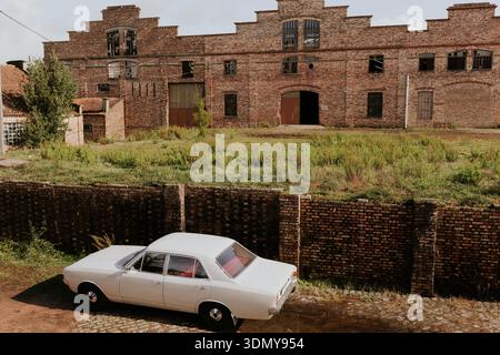 A classic white vintage car is parked on a cobblestone path beside a lush, green field in front of an old, red brick factory. The factory showcases we Stock Photo