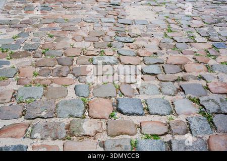 Weathered cobblestones laid in pattern form uneven historic street surface with grass growing between stones Stock Photo