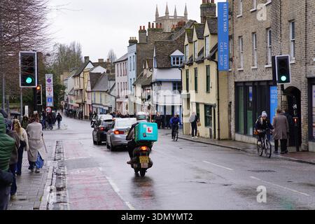 Delivery scooter rider on wet historic street in Cambridge city centre with colourful buildings and pedestrians. Cambridge,England Stock Photo
