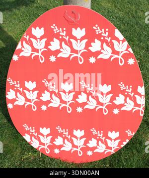 Large red Easter egg decoration with white floral folk pattern displayed on green grass in a park. Minimalist seasonal composition copy space. Stock Photo