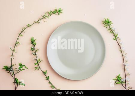Empty ceramic green plate framed by fresh spring branches with white blossoms on soft pastel background. Top view, flat lay. Stock Photo