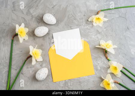 Yellow envelope with blank card surrounded by daffodils and speckled eggs on gray textured background. Happy Easter concept. Top view, flat lay, mocku Stock Photo