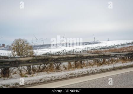 Rows of solar panels stretch across a field dusted with snow, while tall wind turbines turn behind them, showing a mixed renewable energy landscape in Stock Photo