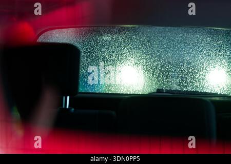 A rear car window is sealed under a rough ice crust, and distant headlights shine into the glass, leaving only a hazy glow and almost no view through Stock Photo