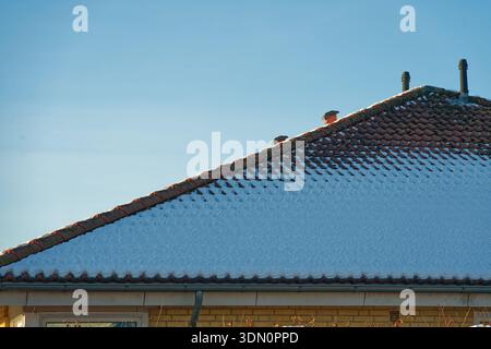 A pinkish tile roof is dusted with snow above a yellow brick house. The light coating highlights the roof pattern and shows a quiet winter morning in Stock Photo