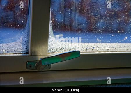 An old wooden window with a worn metal handle is covered in condensation droplets. Cold weather and snow outside create fogging and wet beads on the g Stock Photo