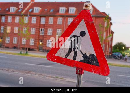 A faded triangular roadworks sign with peeling paint stands near a brick house showing a worker digging symbol and signaling ongoing or past street ma Stock Photo